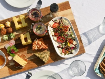 Egy fából készült sajt- és kenőtálca kínálja a Kulináris élvezetek Gasteinben fehér terítőn.