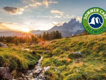 Hegyi táj naplementében, patakkal és fákkal, a sarokban a Filzmoos Sommer Card logójával.