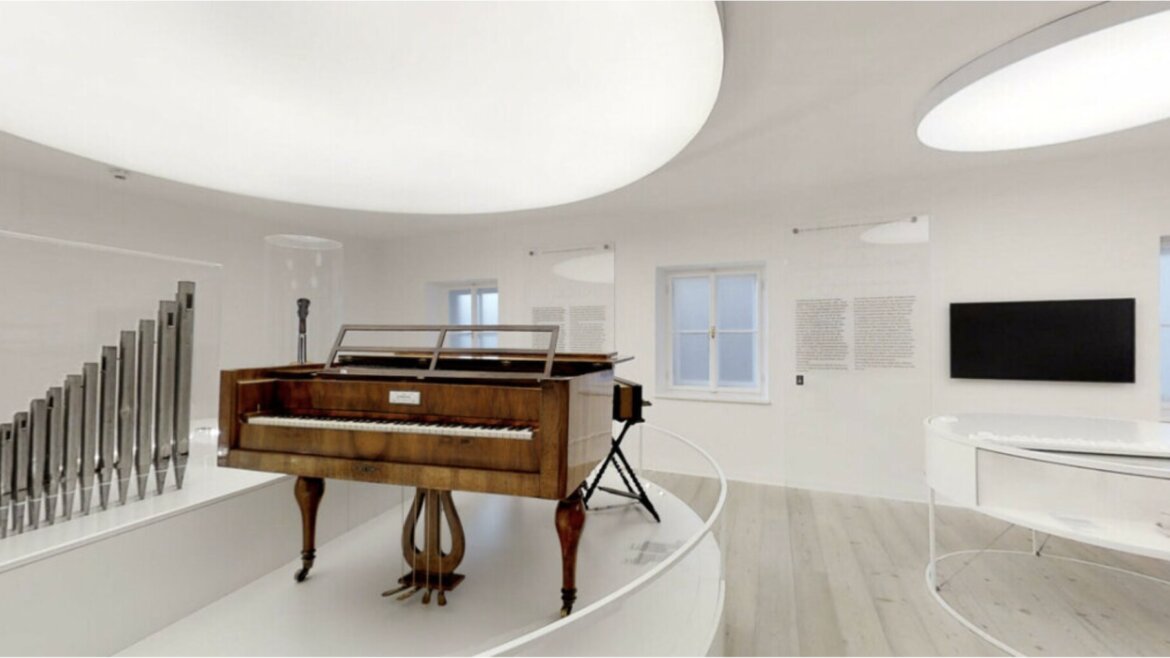 Egy fából készült fortepiano van kiállítva egy világos, modern múzeumi teremben, nagy kerek mennyezeti lámpákkal.