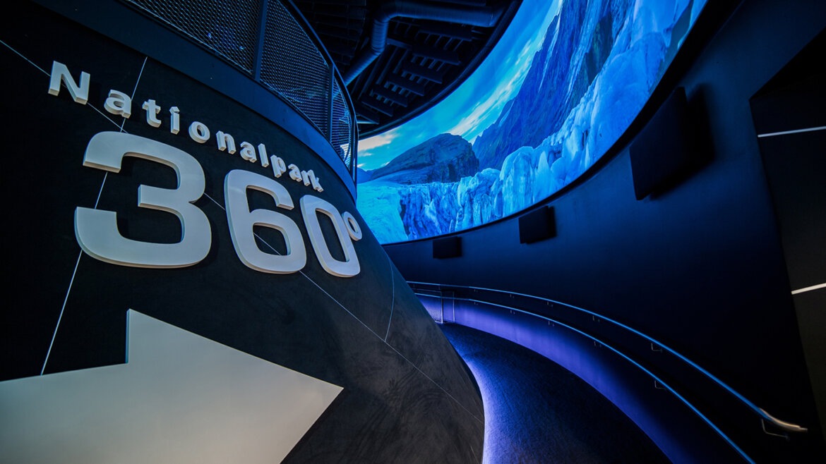 360° Nemzeti Park©Thomas Höll ScienceVision Ívelt folyosó Nationalpark 360° felirattal és egy nagy panorámaképernyővel, amely hegyvidéki tájat mutat. (Nagyított nézet)