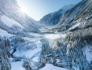 Hóval borított hegyek és erdő, egy folyó kanyarog keresztül, a nap ragyogóan süt a táj felett.