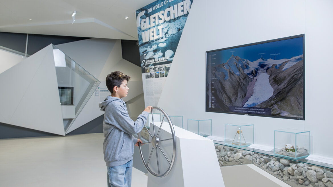Gleccservilág_Fiú© Neumayr/Christian Leopold, www.neumayr.cc, Nationalparkmuseum Egy fiú kereket forgat egy modern gleccsermúzeum gleccserképekkel és vitrinekkel díszített kiállításán. (Nagyított nézet)