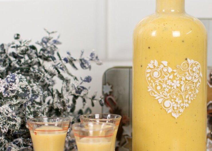 Egy üveg és két pohár krémsárga tojáslikőr áll havas zöld és ünnepi témájú tálcák mellett.