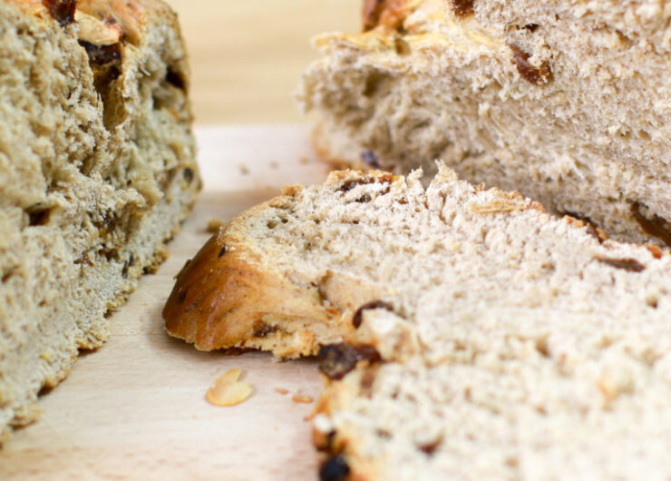 Weihnachtlicher-Striezel-selbst-gemacht-1600×550 Közelkép a szeletelt mazsolás kenyérkenyérről egy fából készült vágódeszkán, a tetején zabpehellyel.