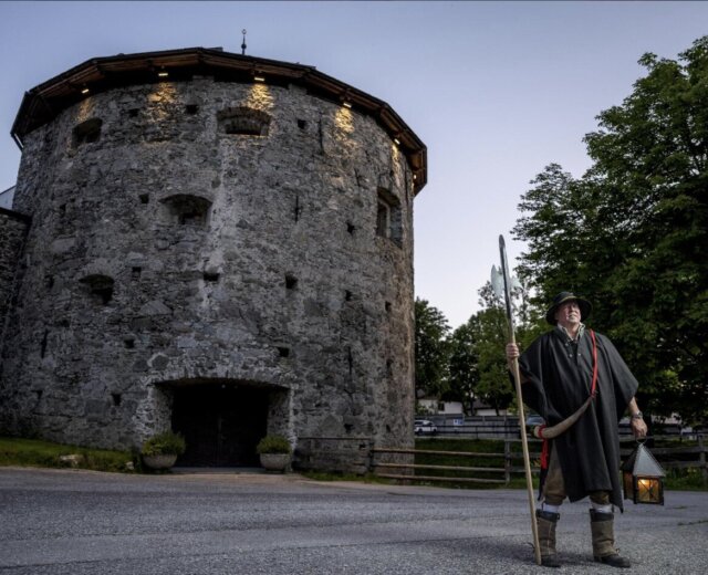 Egy történelmi ruhába öltözött férfi áll botjával és lámpásával egy nagy kőtorony előtt alkonyatkor.