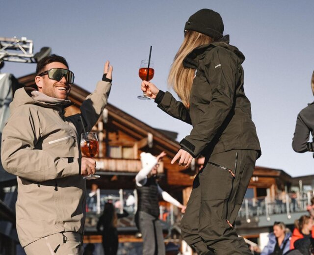 Két sífelszerelésben lévő ember táncol és italozik a szabadban egy fából készült házikó közelében egy napsütéses napon.