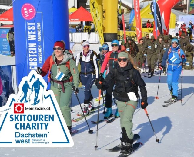 Emberek csoportja síel egy jótékonysági sírendezvényen, mosolyogva és síbotokkal sétálnak színes transzparensek alatt.