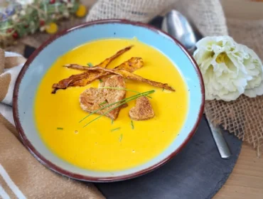 Süßkartoffelsuppe mit Linsen_shared_recipe_1716528615-jpg Egy tál sárga krémleves krutonnal, metélőhagymával és ropogós csíkokkal, mellette egy kanál és egy fehér virág.