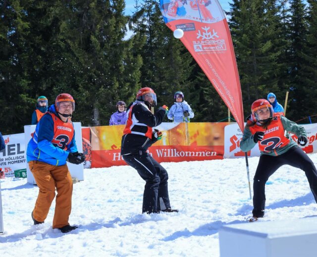 Három piros sisakos ember hógolyókat dobál egy téli versenyen, a háttérben nézők és fák.