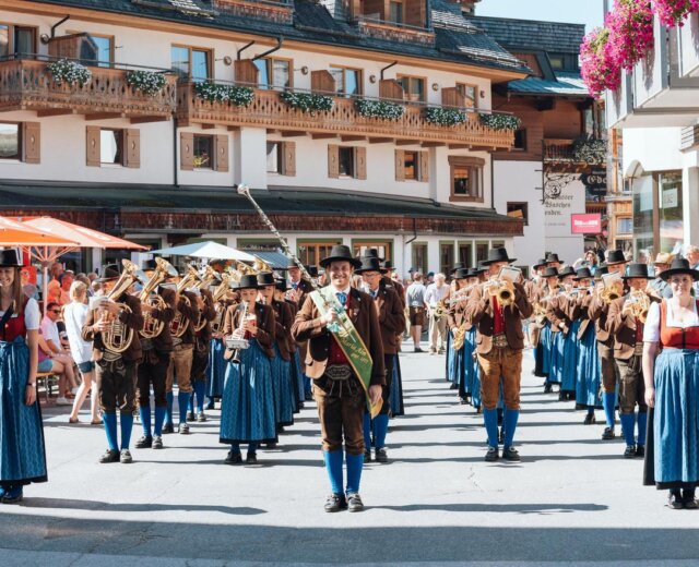 Bauernherbstfest__TMK_Maria_Alm Egy hagyományos, alpesi viseletbe öltözött fúvószenekar a falu főterén ad szabadtéri koncertet, miközben a közelben nézők állnak.