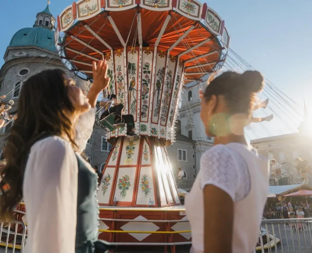karusellfahren-am-rupertikirtag_40969973@2x.ae5b064c Két nő áll egy vidámparkban egy színes hintánál; a háttérben emberek hintáznak, és a napfény ragyog.