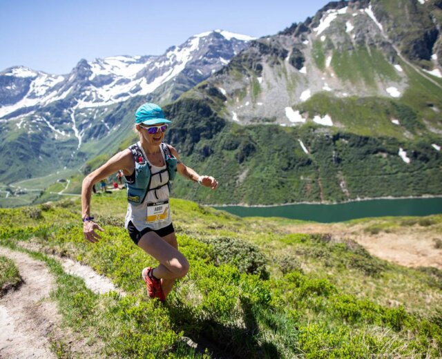 Trailrunning_Adidas_Infinite_Trails Egy sportruhás futó felfelé kocog egy ösvényen, háttérben a hegyekkel és a tóval.