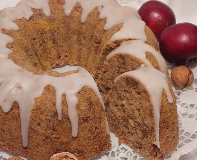 Apfel-Nuss Gugelhupf Cukormázas bundt torta, szeletelve, dióval körülvéve, két piros szilvával egy csipkés szalvétán.