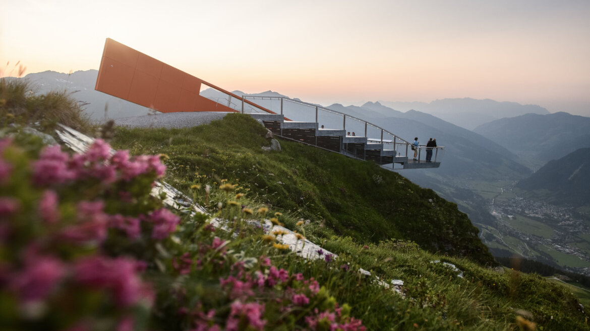 Moderno punto panoramico di montagna con persone, affacciato su una valle al tramonto, con fiori in primo piano.