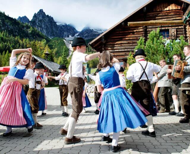 Bambini in abiti tradizionali austriaci ballano all'aperto, con le montagne e una capanna di legno sullo sfondo.