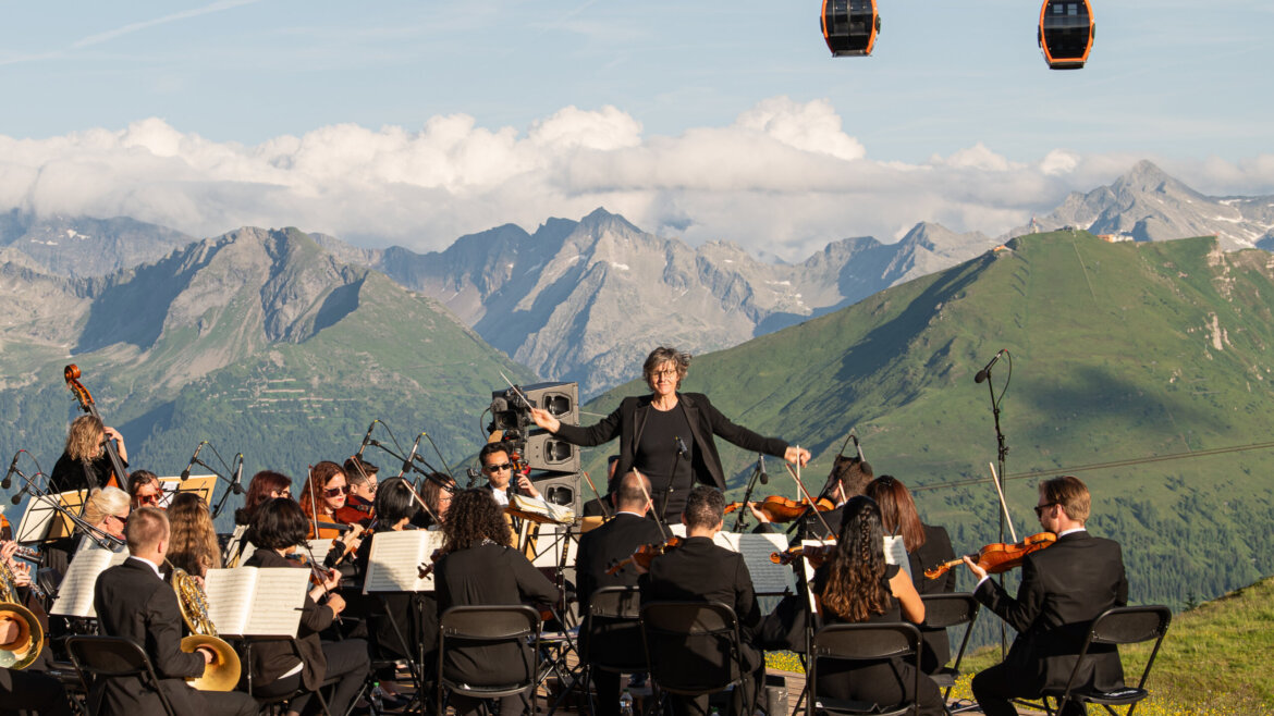 Un'orchestra si esibisce all'aperto su un palco di legno, con montagne e funivie sullo sfondo. (Vista ingrandita)
