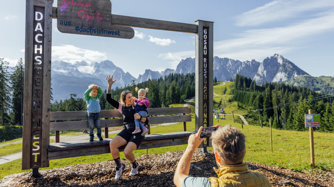 Un uomo scatta una foto di una donna e due bambini su una grande panchina in Tennengau, con le montagne alle spalle. (Vista ingrandita)