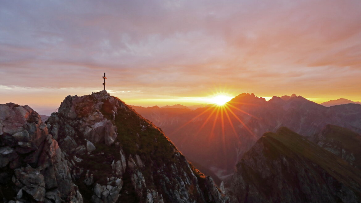 Una croce si erge su un picco roccioso al sorgere del sole, che si affaccia sulle catene montuose del Grossarl illuminate dal sole. (Vista ingrandita)