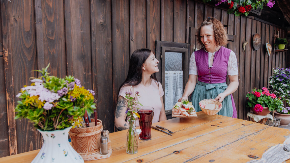 Una donna sorridente serve del cibo a un'altra in un tavolo di legno all'aperto del Tennengau, decorato con fiori. (Vista ingrandita)