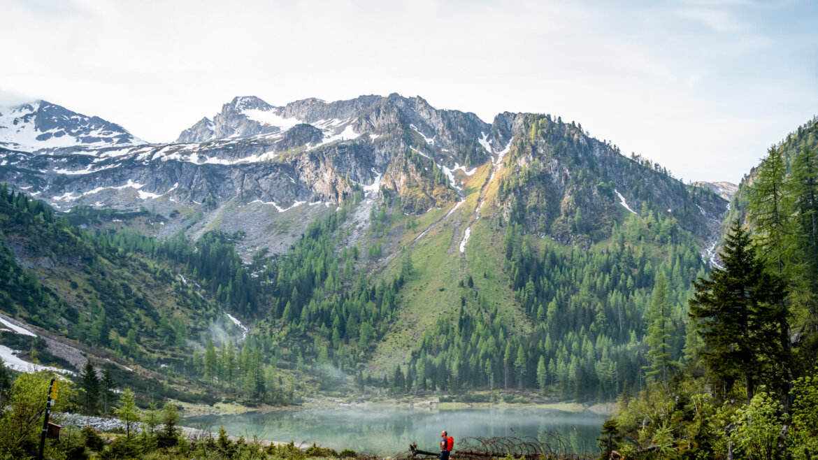 Una persona con una giacca rossa si trova in riva a un lago vicino a Grossarl, con montagne e foreste sotto un cielo blu. (Vista ingrandita)
