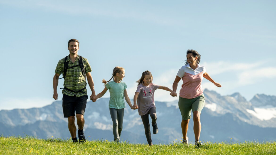 Una famiglia di quattro persone si tiene per mano e corre sull'erba a Grossarl con le montagne sullo sfondo in una giornata di sole. (Vista ingrandita)