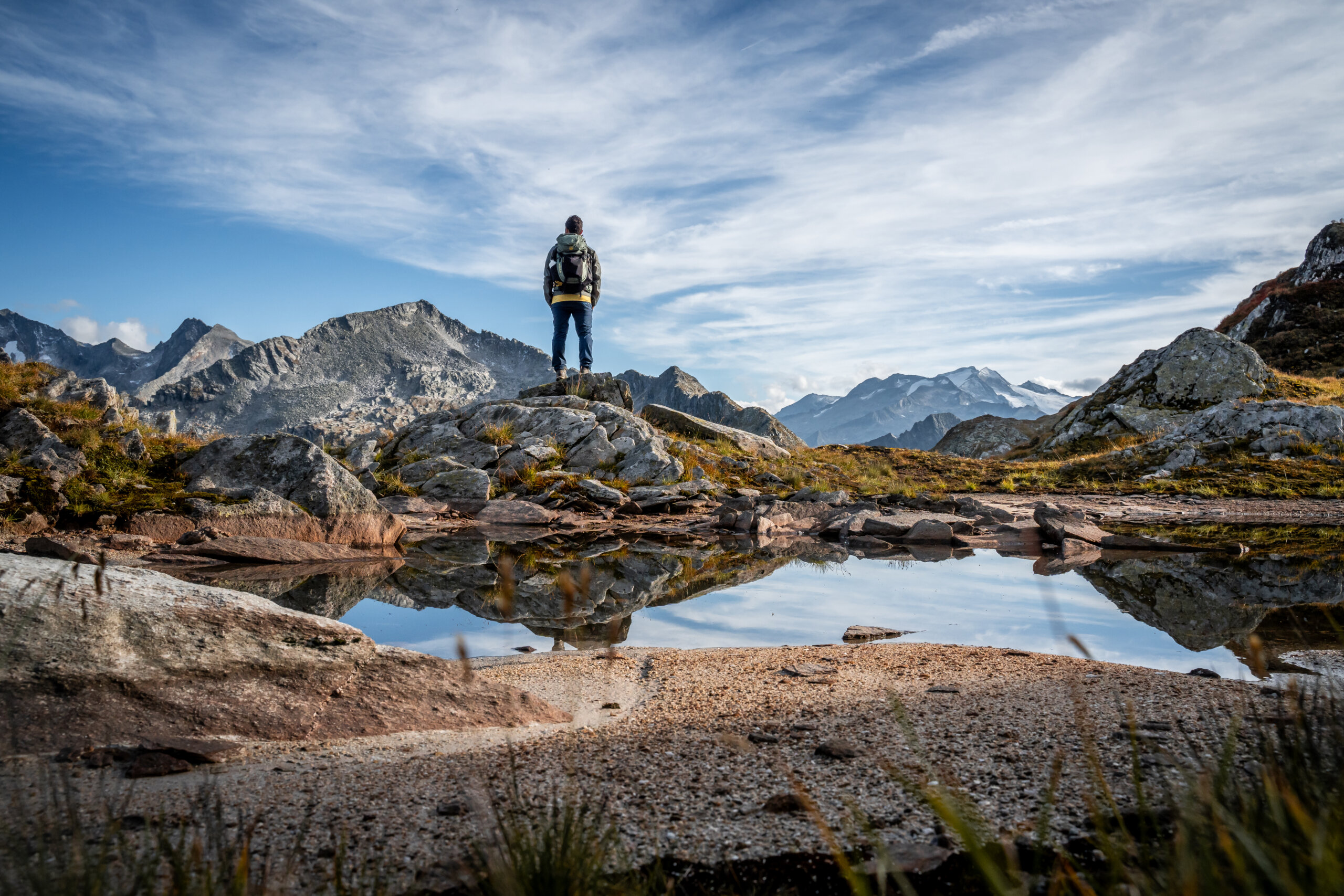 grossarltal-weinschnabel-albert-biwak-mureck-arlscharte-schoedersee Un escursionista si trova su una collina rocciosa a Grossarl, con vista sulle montagne e su uno stagno che riflette il cielo e le cime.