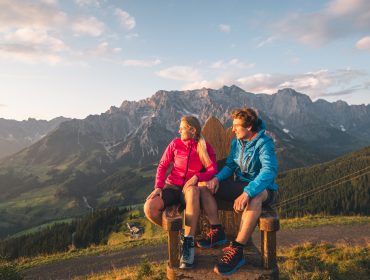 Una coppia in giacca chiara siede su una panchina, con vista sulle montagne al tramonto, nella Zona turistica Hochkönig.