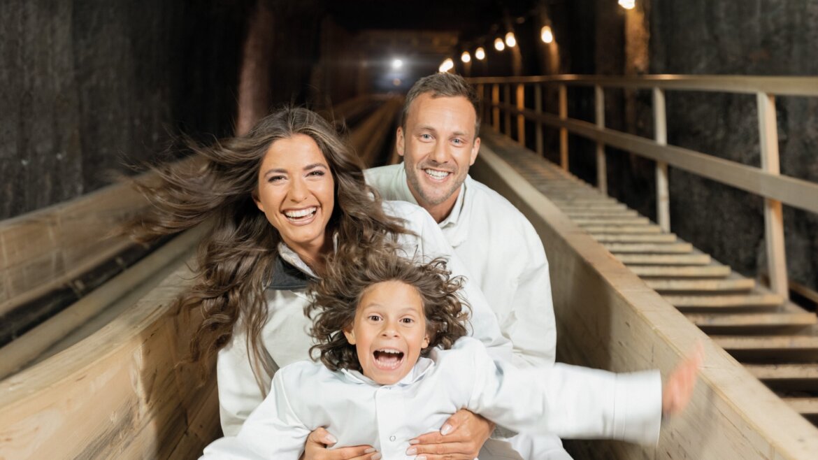 Una famiglia sorridente in abito bianco sale su uno scivolo di legno all'interno di un tunnel del Tennengau poco illuminato. (Vista ingrandita)