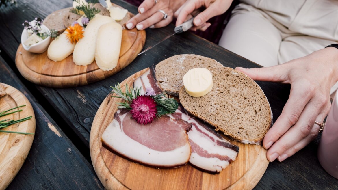 Pane a fette, burro e salumi su un piatto di legno, con una mano che tiene il pane accanto a un altro piatto di formaggio. (Vista ingrandita)