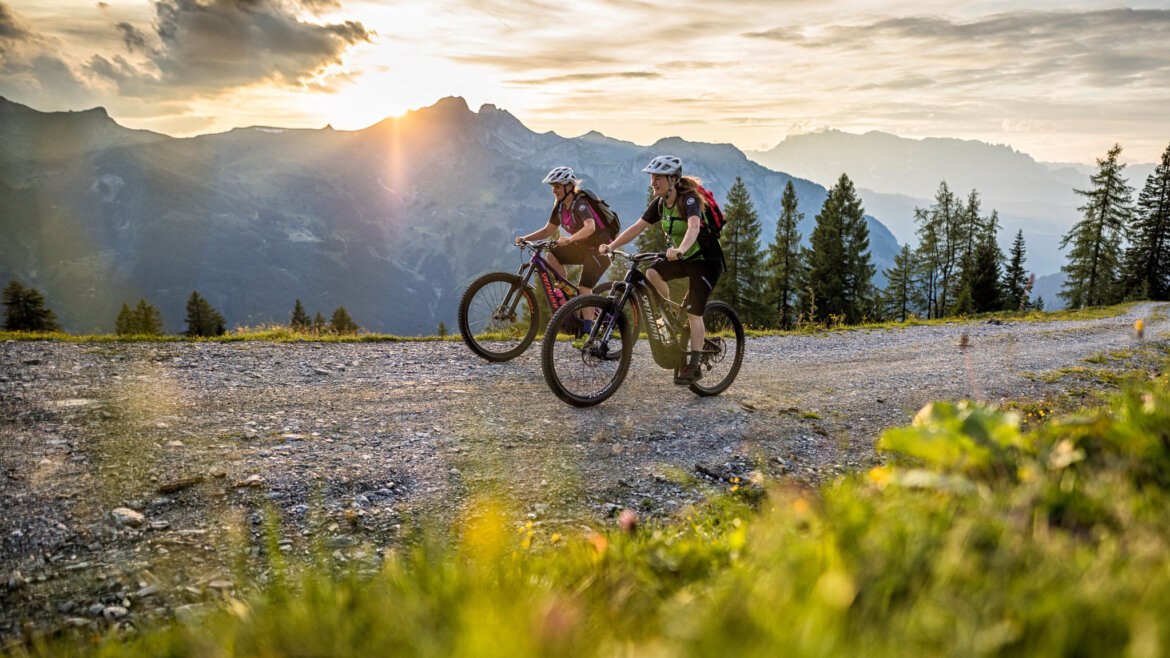 Due persone pedalano in mountain bike su un sentiero di ghiaia a Grossarl al tramonto, circondati da montagne e alberi. (Vista ingrandita)