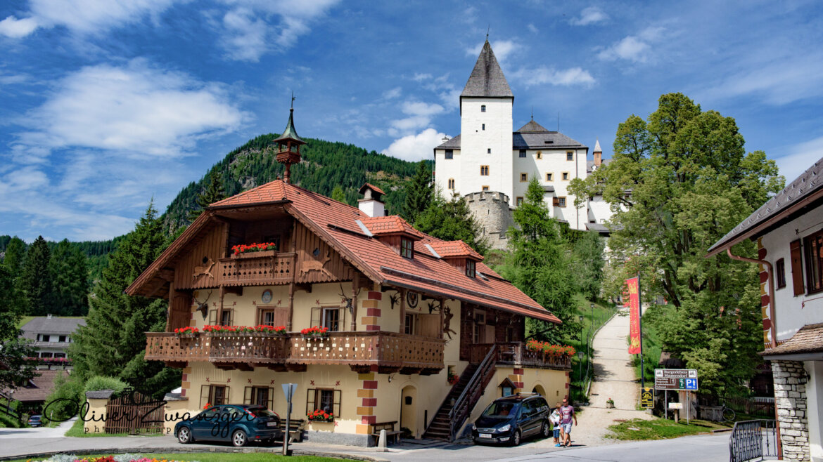 Un edificio in stile chalet nella Zona turistica Lungau, con balconi, un castello in cima alla collina e auto parcheggiate al sole.