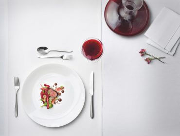 Un piatto gourmet con eccellenze gastronomiche, con vino rosso e un piccolo fiore su una tovaglia bianca.