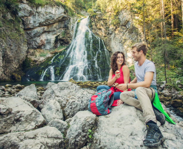 Una coppia sorridente siede su delle rocce vicino a una cascata nella foresta del Tennengau, con gli zaini accanto.