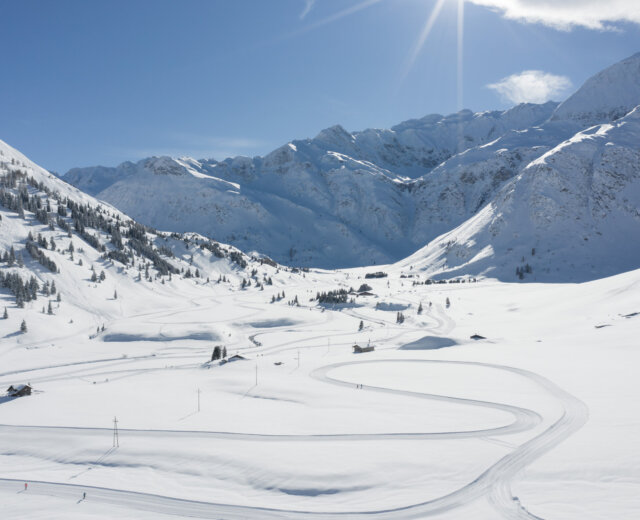 Ski Classics Drohnenaufnahme Loipe Sportgastein; ©Gasteinertal Tourismus GmbH, Christoph Oberschneider Paesaggio montano innevato con piste da sci tortuose, alberi sparsi e luce solare intensa in una giornata limpida.