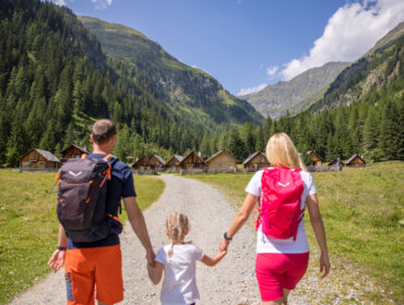 Una famiglia di tre persone percorre un sentiero verso le baite della Zona turistica Lungau, in una rigogliosa valle di montagna.