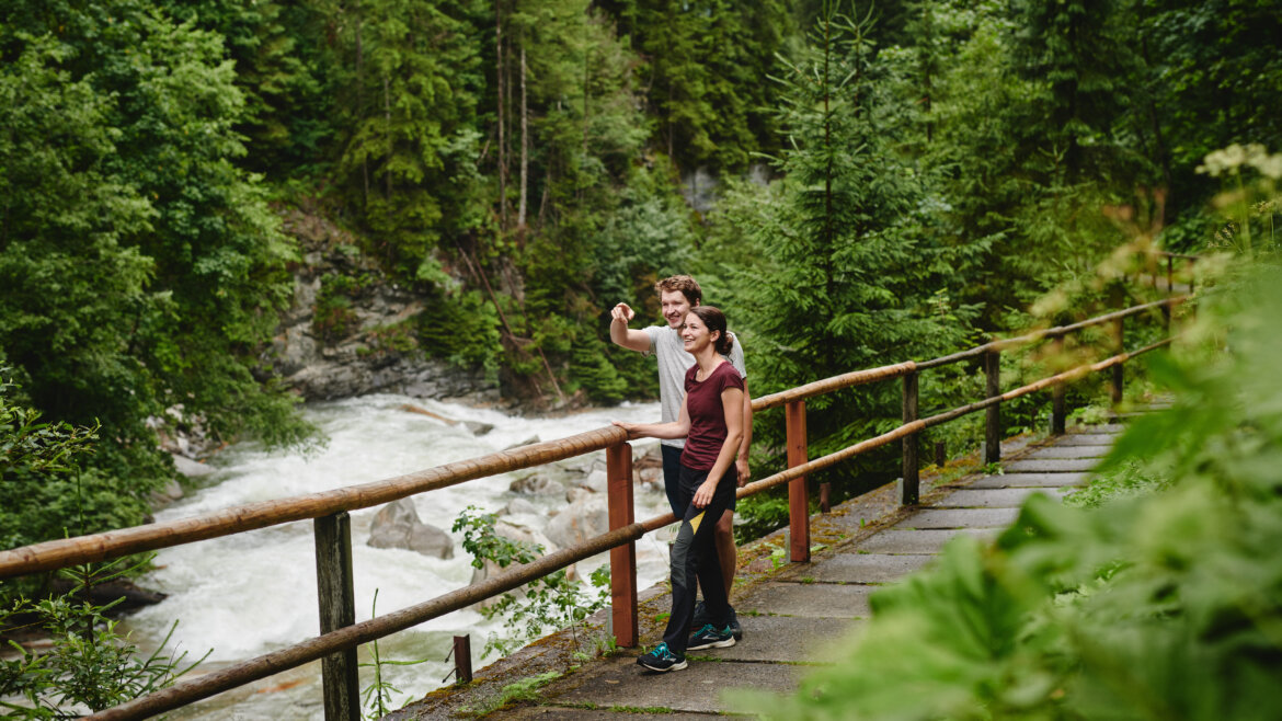 Sentiero delle cascate a Bad Gastein; ©Gasteinertal Tourismus GmbH, Michael Koenigshofer Due persone in piedi su un sentiero lungo il fiume, sorridono e si scattano un selfie circondati da alberi verdeggianti. (Vista ingrandita)