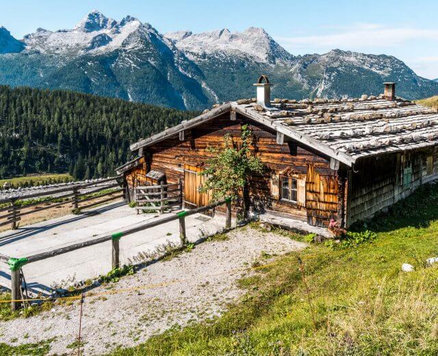 Una rustica capanna di legno si trova su un pendio erboso con le cime delle montagne sullo sfondo.