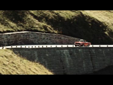 Un'auto d'epoca rossa percorre una tortuosa strada di montagna delimitata da muri di pietra e colline verdeggianti.