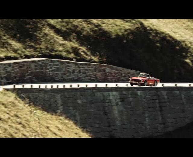 Un'auto d'epoca rossa percorre una tortuosa strada di montagna delimitata da muri di pietra e colline verdeggianti.