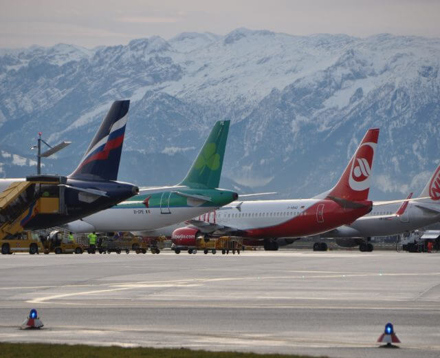 Diversi aerei parcheggiati sulla pista di un aeroporto con montagne innevate sullo sfondo.