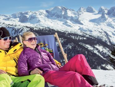 Due persone in abbigliamento da sci colorato si rilassano su sedie nella neve con montagne innevate sullo sfondo.