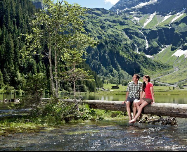 Una coppia siede su un tronco sopra un ruscello limpido con montagne verdi e macchie di neve sullo sfondo.
