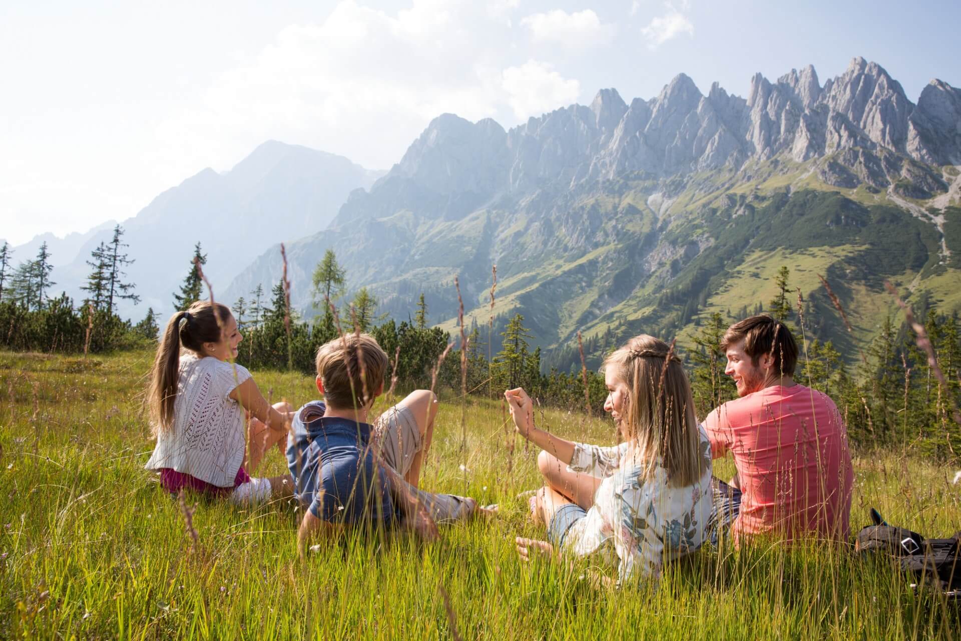 Quattro persone siedono sull'erba in un prato, godendosi la vista delle montagne in una giornata di sole.