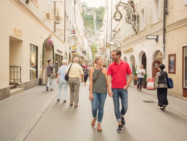 Coppia che si tiene per mano, passeggiando in una vivace via dello shopping salisburghese, fiancheggiata da negozi e pedoni.