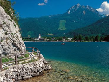 wolgagnseeregion_buergl_strobl_sommer Lago limpido con riva rocciosa, ringhiera in legno, villaggio lontano e sfondo montuoso sotto un cielo blu.