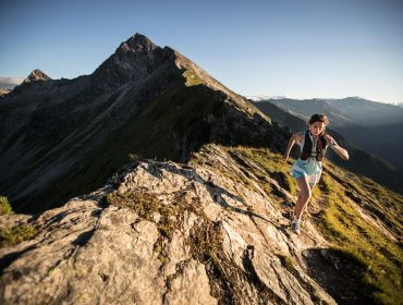 ait_bc_ss_18_michael_mueller__12629 Donna che fa trail running su una cresta rocciosa di montagna con cime e cielo limpido sullo sfondo.