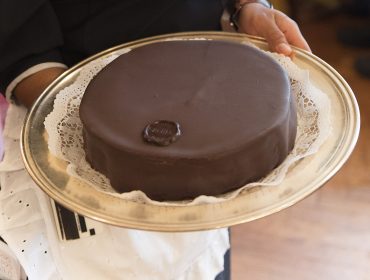 Una persona che tiene in mano un vassoio con una torta rotonda ricoperta di cioccolato su un centrino di pizzo.