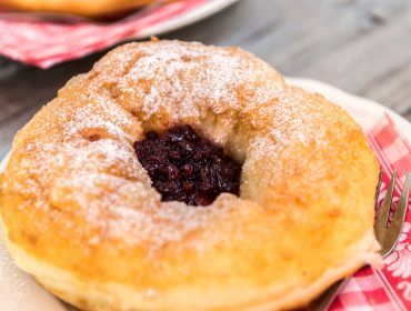 Bauernkrapfen mit Preiselbeeren Una pasta fritta con zucchero a velo e marmellata di frutti di bosco al centro, servita su un piatto con tovagliolo rosso e forchetta.