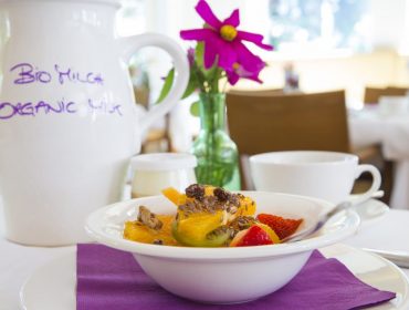 bioparadies_auersperg_frueh-1170×658 Ciotola di frutta fresca e granola su un tavolo, con cibo genuino, brocca di latte e vaso di fiori rosa.