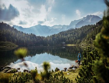 Yoga sul lago Reedsee Persona che pratica yoga presso un tranquillo lago di montagna, circondato da alberi di legno e da lontane cime innevate.